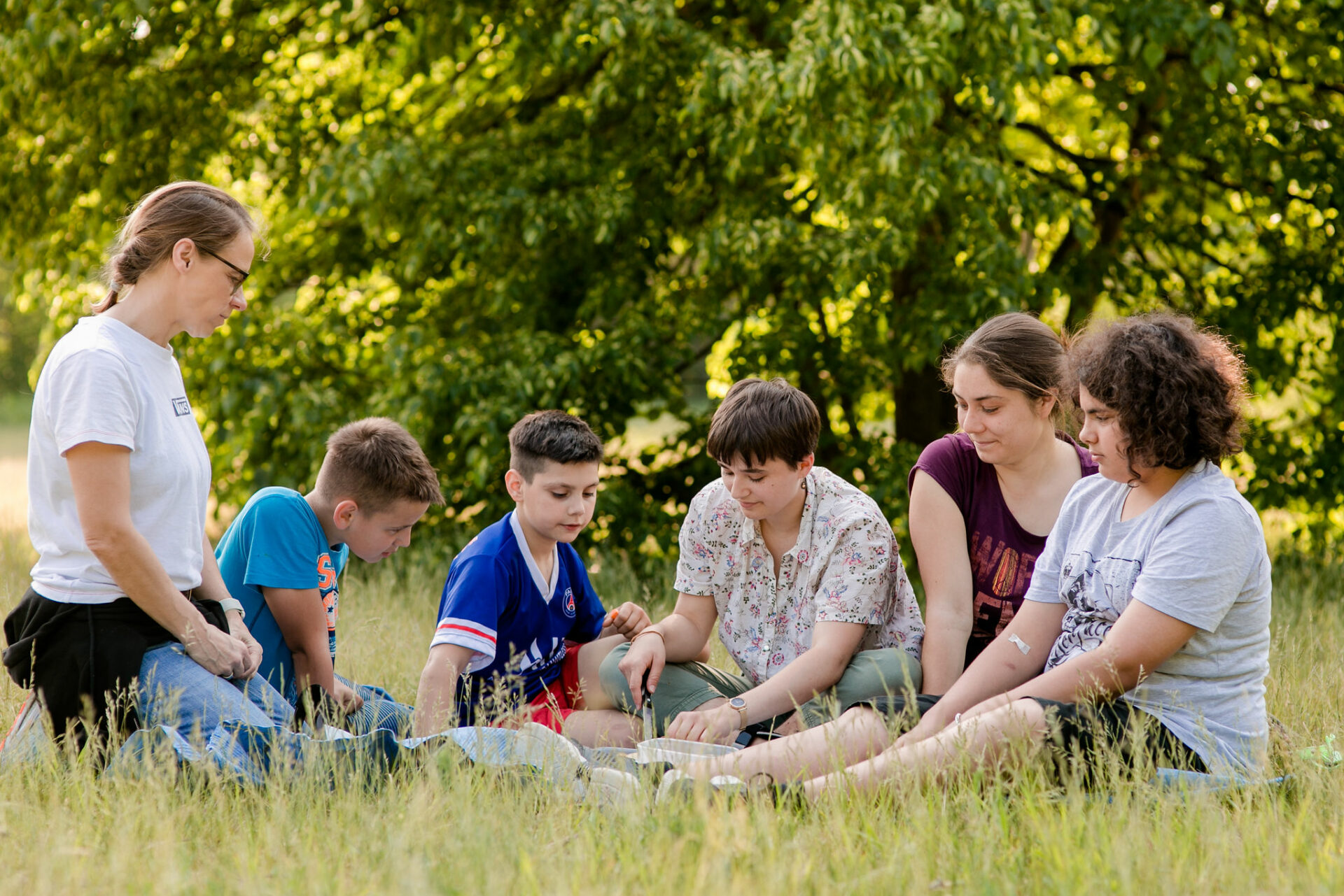 Teenager und Mitarbeitende von Stoffwechsel Dresden-Pieschen erleben Gemeinschaft beim gemeinsamen Picknick auf einer grünen Wiese mit Bäumen im Hintergrund