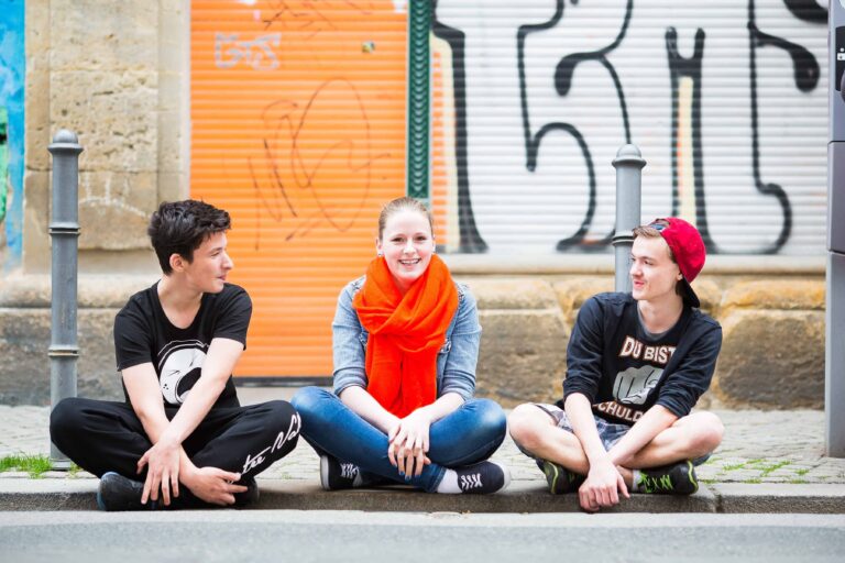 Drei Teenager sitzen auf dem Bordstein vor m Hauptstandort des Stoffwechsel e.V. - mit Streetart Graffiti an der Hauswand im Hintergrund.
