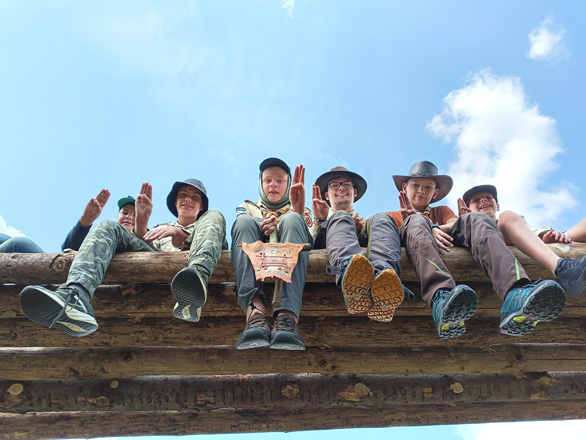 Kinder und Jugendliche in Pfadfinderkluft und Hut sitzen auf Holzbalken und zeigen das Pfadfinder-Zeichen der Royal Rangers mit der Hand. Einer hält ein kleines Banner mit der Aufschrift Sommercamp vor sich.